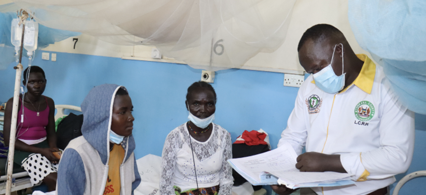 A doctor, standing and looking over a chart, attends to a patient seated on a cot.
